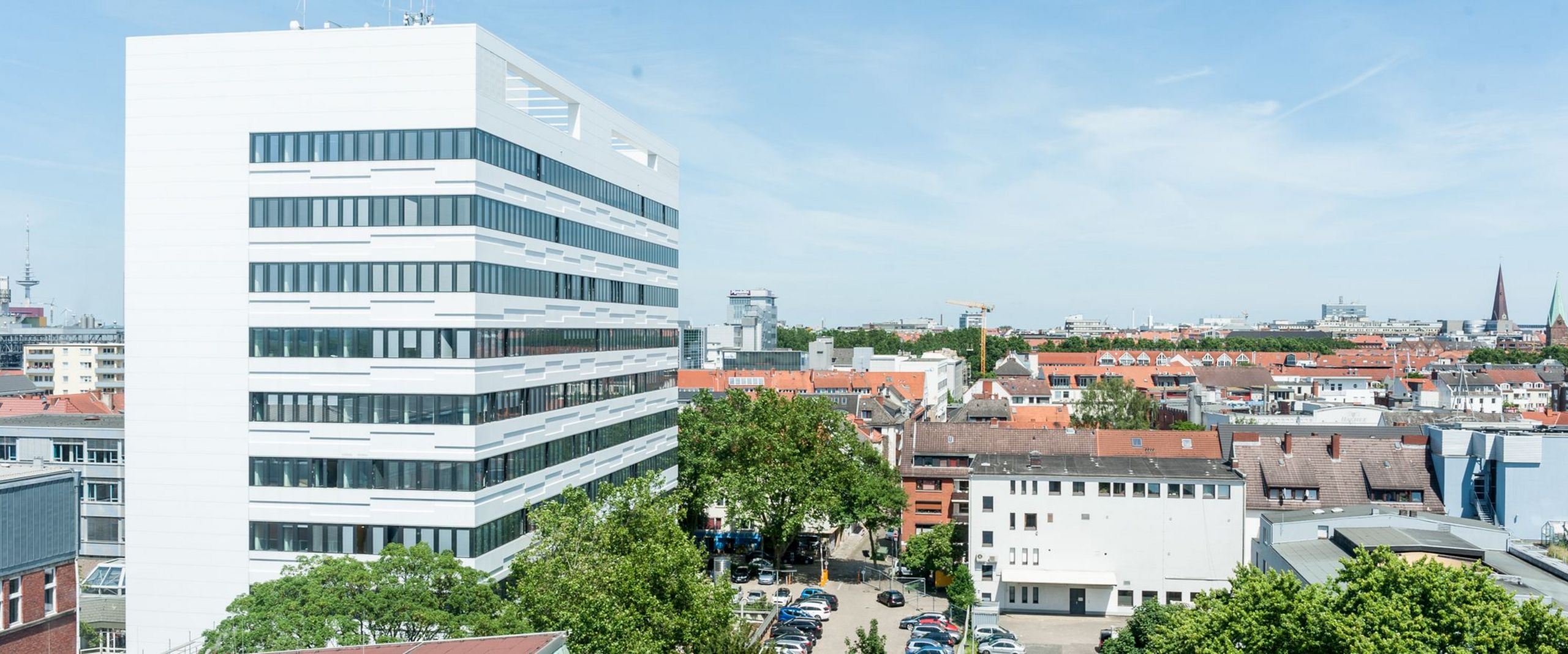 Auf dem Bild ist das AB-Gebäude des Campus Neustadtswall zusehen. Das Foto ist aus der Luft aufgenommen, sodass man außerdem noch blauen Himmel, grüne Bäume und einige Nebengebäude und den Parkplatz erkennen kann. 